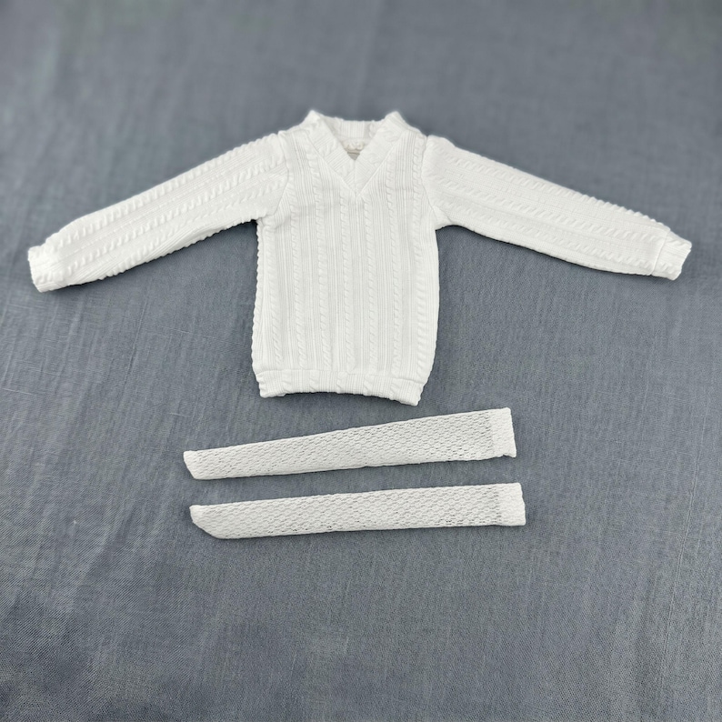 Puede incluir: Un su&eacute;ter blanco de manga larga con cuello en V y un patr&oacute;n de tejido de cable vertical, acompa&ntilde;ado por dos piezas de tela rectangulares blancas separadas con un dise&ntilde;o de malla de tejido abierto texturizado colocado debajo del su&eacute;ter.