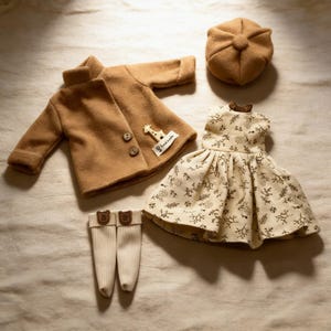 K&ouml;nnte beinhalten: Ein beiges Puppenkleidungsset mit einem Mantel mit Holzkn&ouml;pfen, einem Kleid mit floralem Muster, einer M&uuml;tze und gestreiften Socken mit B&auml;renakzenten. Der Mantel hat eine Giraffenapplikation.