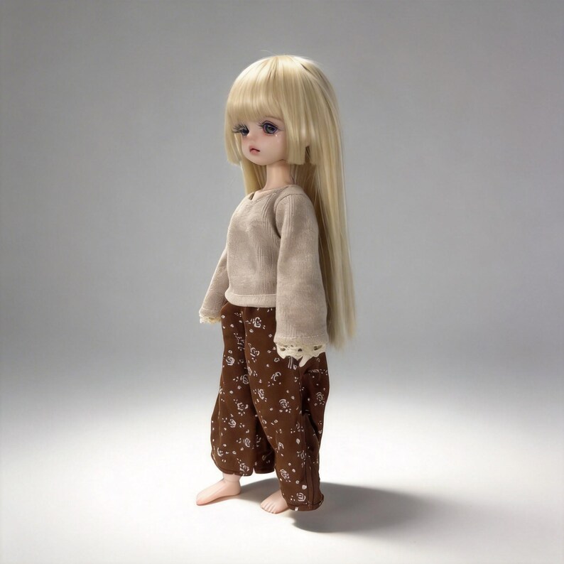 May include: A doll with long, straight blonde hair and a neutral expression. The doll wears a beige long-sleeved shirt with lace trim and brown pants with a white floral pattern. The doll is barefoot and stands against a plain background.