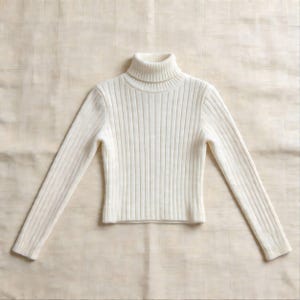 May include: A cream-colored turtleneck sweater with long sleeves. The sweater has a ribbed knit design and is made of a soft, warm material. The turtleneck is folded over and the sleeves are extended.