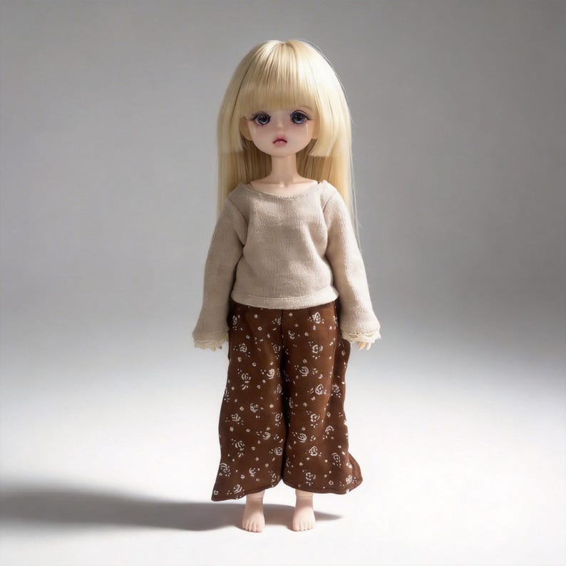 May include: A doll with long, straight blonde hair and a neutral expression. The doll wears a beige long-sleeved shirt and brown, wide-legged pants with a white floral pattern. The doll's eyes are a light purple.