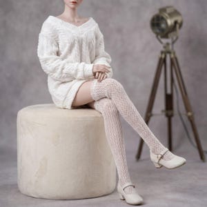 Puede incluir: Una mu&ntilde;eca viste un su&eacute;ter blanco de cuello en V con tejido de ochos, medias blancas texturizadas por encima de la rodilla y zapatos Mary Jane blancos con una peque&ntilde;a correa de hebilla. La mu&ntilde;eca est&aacute; sentada, mostrando el tejido.