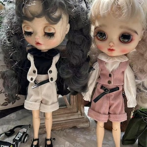 May include: Two fashion dolls with large eyes and long eyelashes. One doll wears a black top, cream-colored vest and shorts, and black shoes. The other doll wears a pink vest and shorts, a white blouse, and black shoes. Both dolls have long hair.