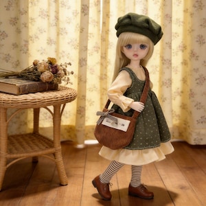 May include: A doll wearing a green dress with white polka dots, a matching hat, and a brown corduroy bag. The doll has blonde hair, blue eyes, and is wearing brown shoes and striped socks. A small table with a book and flowers is in the background.