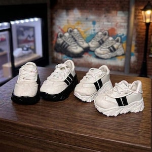 May include: Four miniature sneakers arranged on a wooden surface. Each shoe is white with black side stripes, thick white soles, and white laces. The design features a textured upper material and a chunky silhouette, mimicking the appearance of athletic footwear.