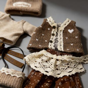 May include: A collection of doll clothes in neutral tones. The ensemble includes a brown knit vest with white heart accents, a cream-colored lace collar, and brown pants with a white dot pattern. A small purse with a lace trim and a knit hat are also visible.