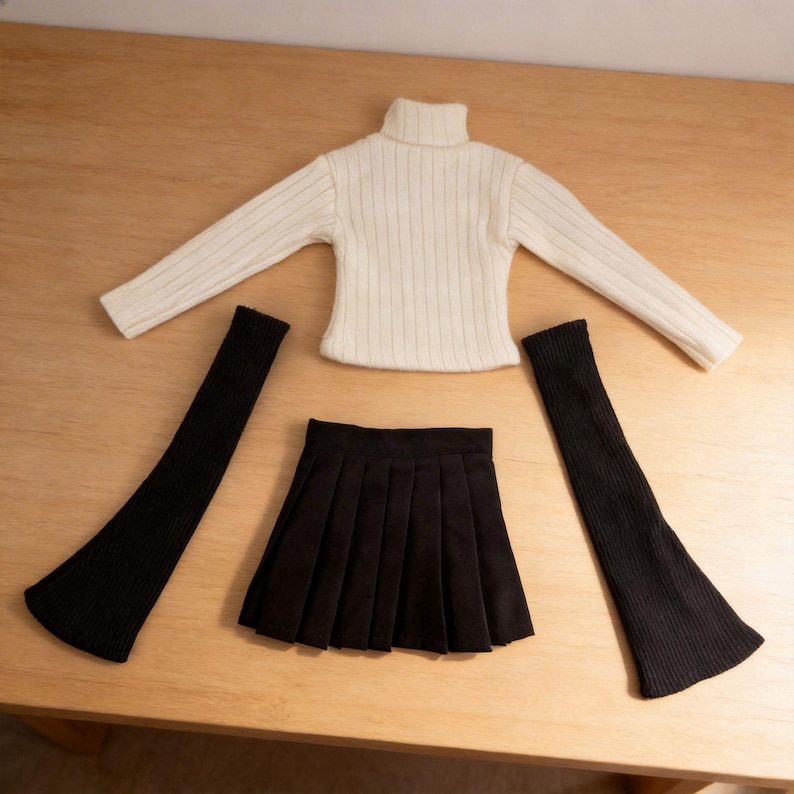 May include: A doll-sized outfit set featuring a cream-colored ribbed turtleneck sweater, a black pleated mini skirt, and two black arm warmers. The clothing items are arranged on a light wooden surface.