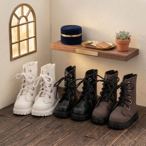 May include: Three pairs of miniature lace-up boots in white, black, and grey, each featuring silver-toned eyelets, thick black or white laces, and chunky black lug soles. The boots are arranged in a row on a dark wooden floor surface.