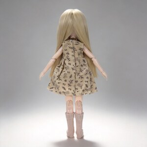 K&ouml;nnte beinhalten: Eine Puppe mit langen blonden Haaren, die ein beiges Kleid mit schwarzem Blumenmuster tr&auml;gt. Die Puppe hat bewegliche Gelenke und tr&auml;gt rosa kniehohe Socken. Die Puppe steht mit dem R&uuml;cken zum Betrachter.