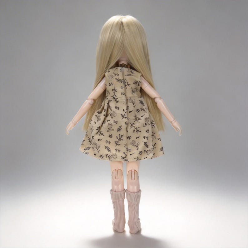 K&ouml;nnte beinhalten: Eine Puppe mit langen blonden Haaren, die ein beiges Kleid mit schwarzem Blumenmuster tr&auml;gt. Die Puppe hat bewegliche Gelenke und tr&auml;gt rosa kniehohe Socken. Die Puppe steht mit dem R&uuml;cken zum Betrachter.