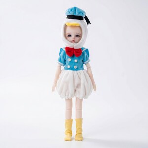 Op de afbeelding: Een pop verkleed als Donald Duck. De pop draagt een blauw matrozenhemd met parelknopen, een witte rok en gele laarzen. Een blauwe matrozenhoed en een rode vlinderdas maken de outfit compleet. De pop heeft een witte kap met een gele snavel.