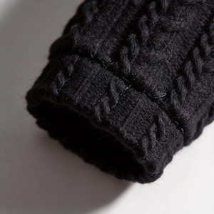 Pu&ograve; includere: Primo piano di un polsino di manica in maglia nera. Il polsino presenta un motivo a trecce, con punti spessi e strutturati. Il colore scuro e il motivo dettagliato creano un aspetto accogliente e tattile. La manica fa probabilmente parte di un maglione o di una giacca.