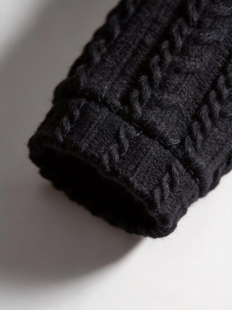 Pu&ograve; includere: Primo piano di un polsino di manica in maglia nera. Il polsino presenta un motivo a trecce, con punti spessi e strutturati. Il colore scuro e il motivo dettagliato creano un aspetto accogliente e tattile. La manica fa probabilmente parte di un maglione o di una giacca.