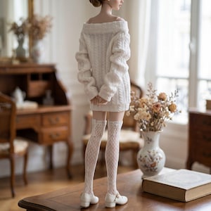 Puede incluir: Una mu&ntilde;eca viste un su&eacute;ter blanco de punto trenzado con hombros descubiertos, medias blancas de encaje hasta el muslo y zapatos blancos de tac&oacute;n bajo. La mu&ntilde;eca se ve desde atr&aacute;s, mostrando el patr&oacute;n de punto y el dise&ntilde;o de encaje.