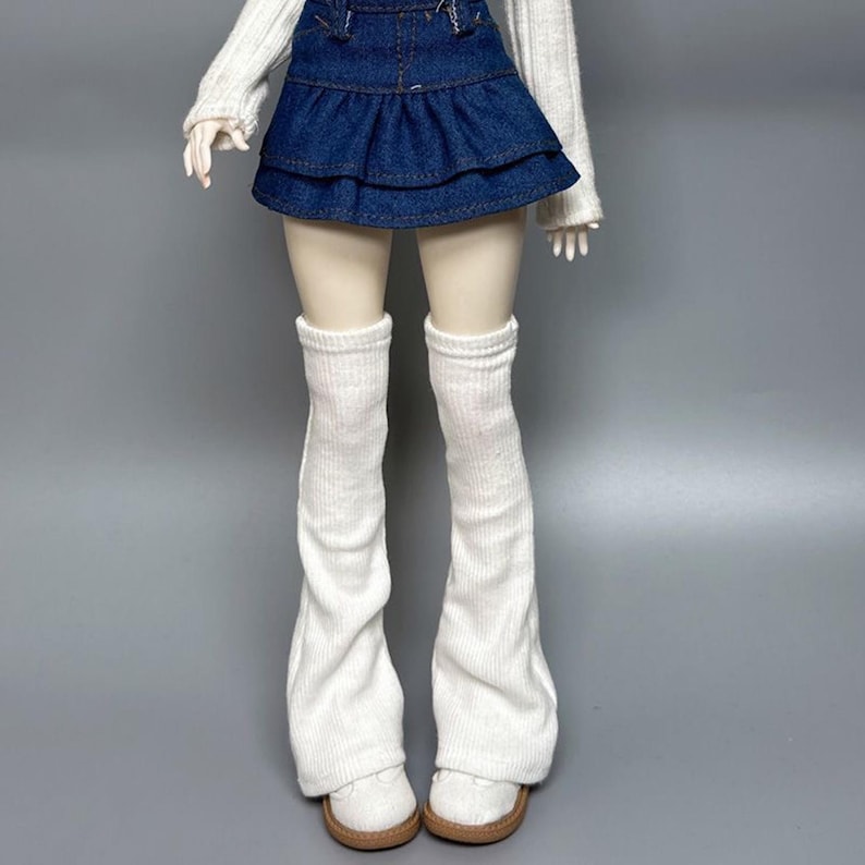 May include: A doll wearing a white sweater, a denim skirt, white leg warmers, and white shoes with brown soles. The doll is posed against a gray background. The outfit is a casual style.