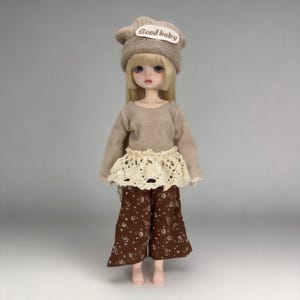 May include: A doll wearing a beige knit cap with "Good baby" text, a long-sleeved beige top, a cream-colored lace skirt, and brown floral print pants. The doll has long blonde hair and is standing.