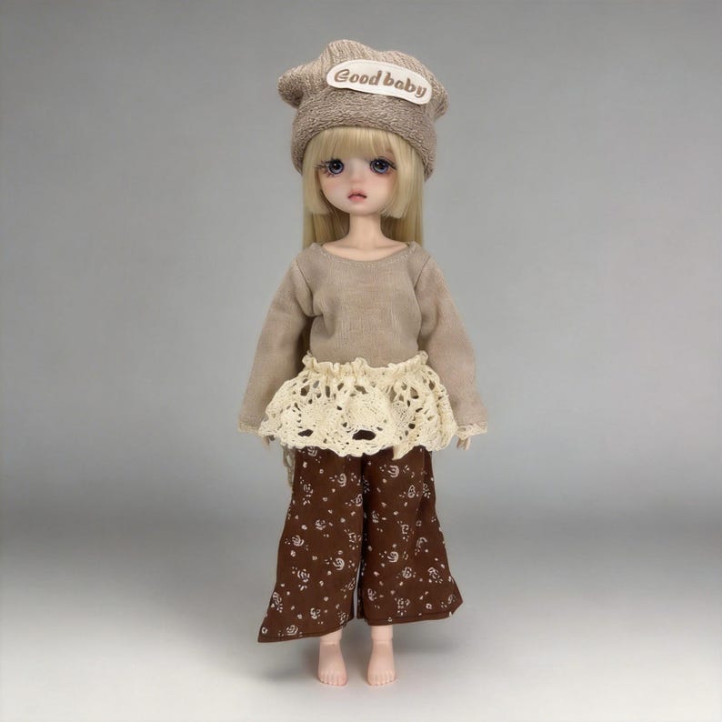 May include: A doll wearing a beige knit cap with "Good baby" text, a long-sleeved beige top, a cream-colored lace skirt, and brown floral print pants. The doll has long blonde hair and is standing.