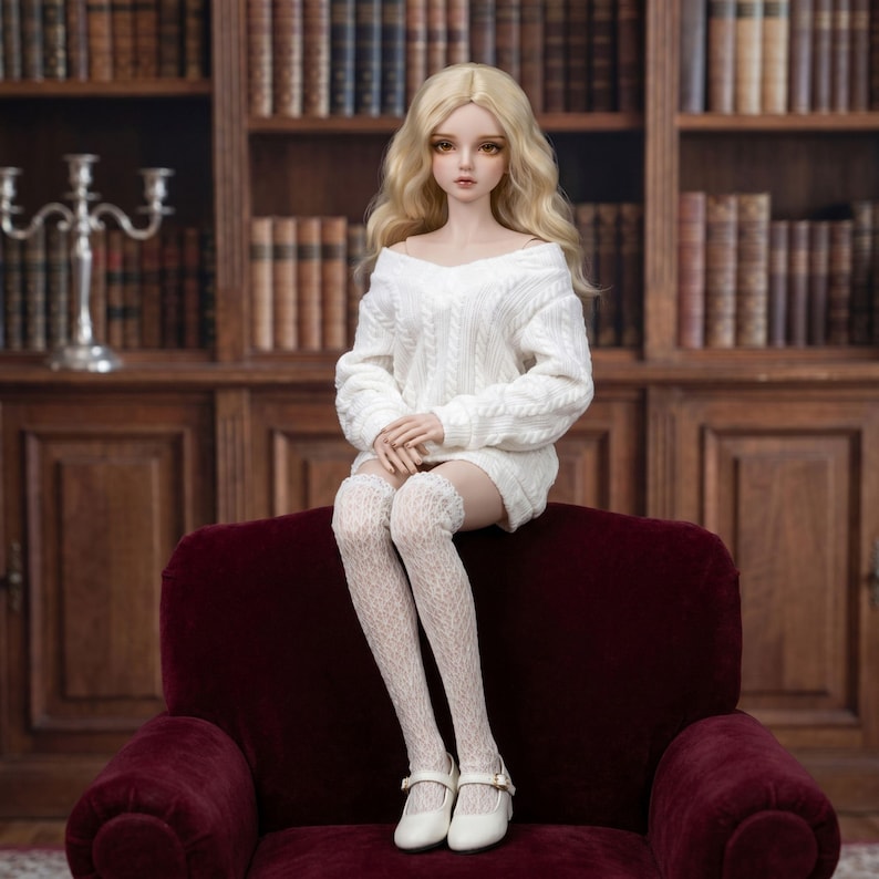 Puede incluir: Una mu&ntilde;eca articulada con cabello largo rubio, ojos marrones y tez p&aacute;lida. La mu&ntilde;eca viste un su&eacute;ter blanco de punto trenzado, medias blancas de encaje hasta el muslo y zapatos Mary Jane blancos con peque&ntilde;as hebillas, en posici&oacute;n sentada.