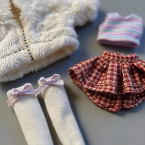 May include: A doll clothing set featuring a white fuzzy jacket, a striped top, a red and white checkered skirt, and white socks with pink bows. The miniature outfit is designed for a doll.