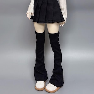 May include: A doll wearing a black pleated skirt, a white sweater, and black corduroy leg warmers. The doll is also wearing white shoes with tan soles. The background is a solid gray.