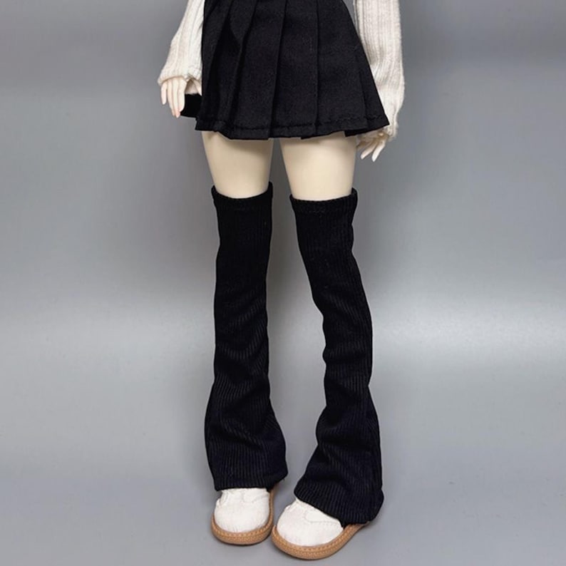 May include: A doll wearing a black pleated skirt, a white sweater, and black corduroy leg warmers. The doll is also wearing white shoes with tan soles. The background is a solid gray.