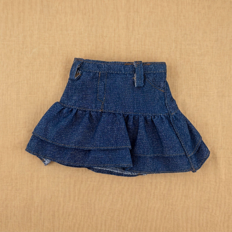 May include: A blue denim skirt with a ruffled hem. The skirt has a classic five-pocket design with a button and belt loops. The denim is a medium wash, and the skirt appears to be new.