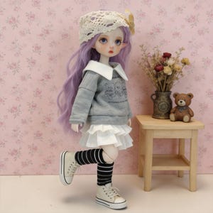 Puede incluir: Una muñeca con cabello lavanda, que lleva una sudadera gris con cuello blanco y una falda blanca. La muñeca también lleva calcetines a rayas blancas y negras y zapatillas blancas. Un sombrero de ganchillo con un lazo amarillo está sobre la cabeza de la muñeca.