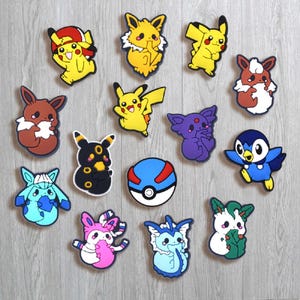 May include: A collection of colorful, cartoon character-shaped magnets. The magnets feature various characters in yellow, brown, purple, blue, and pink. A red and white ball-shaped magnet is also included. The magnets are arranged on a light gray surface.