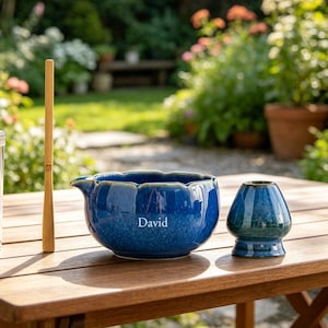 Könnte beinhalten: Ein Matcha-Teeset auf einem Holztisch im Freien. Das Set umfasst eine blaue Keramikschale mit dem Aufdruck "David", einen Bambusbesen in einem transparenten Behälter, einen Bambuslöffel und eine kleine, dunkelblaue Keramik-Teeschale. Im Hintergrund ein Garten.