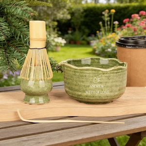 May include: A matcha tea set featuring a green ceramic bowl, a bamboo whisk, and a bamboo scoop. The bowl is inscribed with "ANDREW WITH OF LOVE". A paper coffee cup is also visible.