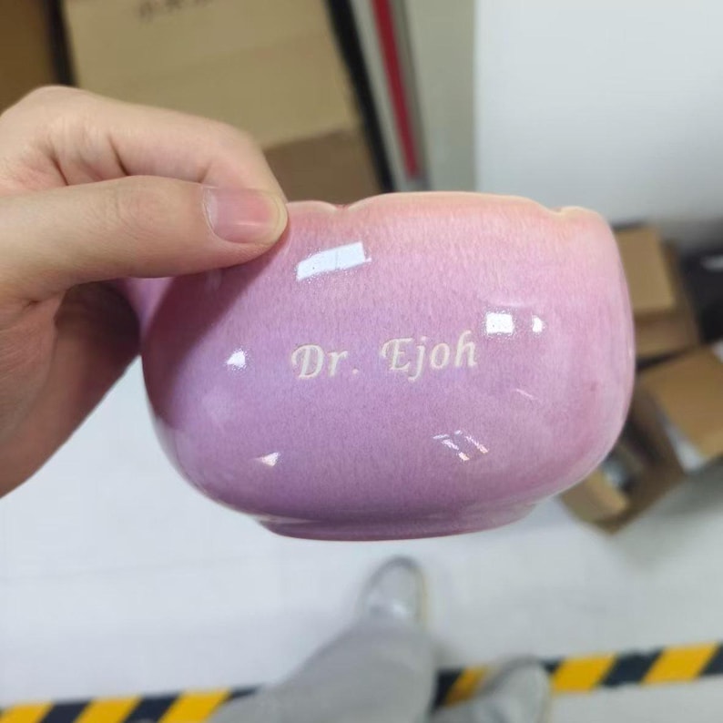 May include: A rounded ceramic mug with a glossy, light pink gradient finish. The surface features the text Dr. Ejoh engraved in a clean, white serif font. The mug has a smooth, curved shape and a reflective surface that catches the light.