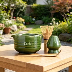 May include: A green ceramic matcha bowl with the name "David" in white, a bamboo whisk, a green matcha bowl, and a wooden scoop on a wooden table. The background shows a garden with greenery and sunlight.