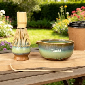 Op de afbeelding: Een matcha-theeset met een bamboe klopper, een keramische kom met de naam "David" en een bamboe lepel. De kom heeft een verloop van bruin, groen en blauw. Een papieren koffiebeker staat op de achtergrond.