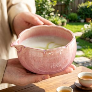May include: A pink ceramic bowl with a scalloped rim and a small pouring spout. The exterior features a glossy, mottled glaze finish with the name Ella written in white cursive script on the side. The bowl contains a light-colored liquid.