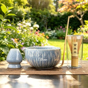 May include: A matcha tea set featuring a blue ceramic bowl with the name "Ethan", a whisk, a whisk holder, and a bamboo scoop. The whisk is in a clear container with Japanese text. The set is on a wooden surface.