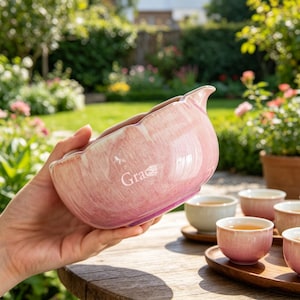 May include: A pink ceramic tea bowl with a scalloped rim and a glossy, marbled finish. The word Grace is printed in white on the side of the bowl. The bowl features a small pouring spout and is held against a blurred outdoor garden background.