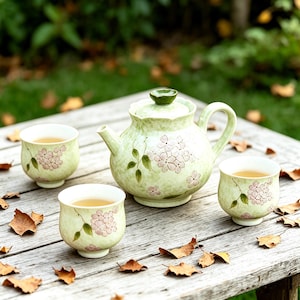 Peut inclure: Un service à thé en céramique vert clair comprenant une théière et deux tasses, chacune ornée de motifs floraux roses et de feuilles vertes. La théière a un couvercle assorti et est posée sur une table en bois vieilli. Les tasses sont remplies de thé.