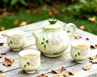 May include: A light green ceramic tea set with a teapot and two teacups, each adorned with pink floral designs and green leaves. The teapot has a matching lid and is placed on a weathered wooden table. The teacups are filled with tea.