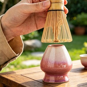 May include: A bamboo matcha whisk with thin, light-colored bristles held above a ceramic matcha bowl. The bowl features a smooth, glossy finish with a gradient transition from a creamy white top to a deep, vibrant pink base with a wide rim.