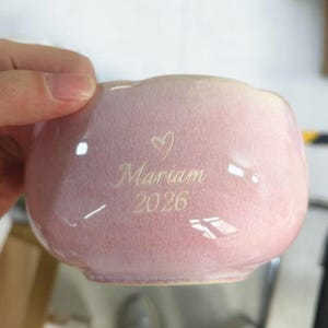 May include: A light pink ceramic bowl with a smooth, glossy finish. The front surface features an engraved design consisting of a small heart symbol above the name Mariam and the year 2026, all centered in a clean, elegant script font.