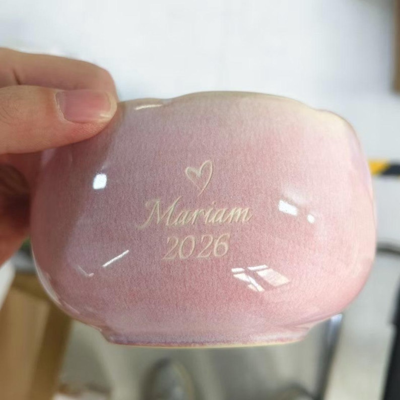 May include: A light pink ceramic bowl with a smooth, glossy finish. The front surface features an engraved design consisting of a small heart symbol above the name Mariam and the year 2026, all centered in a clean, elegant script font.