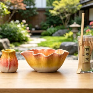 May include: A ceramic tea set in shades of yellow and orange. The set comprises a small vase, a bowl with a wavy rim, and a container holding a bamboo whisk and a label with Japanese text.