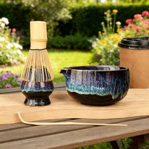 May include: A matcha tea set featuring a bamboo whisk, a ceramic bowl with the name "ETHAN", and a bamboo scoop. The bowl has a blue and black glaze. A paper coffee cup with a black lid is also present.