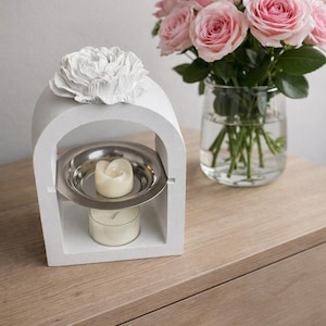 Peut inclure: Un bougeoir blanc en forme d'arche, surmonté d'une fleur, contenant une bougie dans un plat en métal. Un vase de roses roses est posé à côté, sur une surface en bois. Un élément de décoration pour la maison.