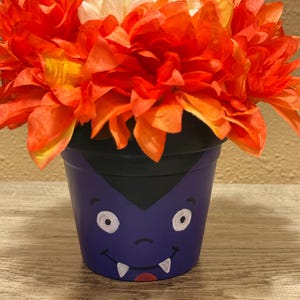 May include: A purple painted flower pot featuring a hand-painted vampire face with white fangs, black hair, and round eyes. The pot is filled with a dense arrangement of vibrant orange and white artificial flowers with layered petals.