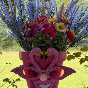 May include: A pink terracotta flower pot featuring a painted face with a wide smile and long eyelashes. The pot is adorned with glittery pink flamingo-shaped glasses and a small pink pom-pom nose, filled with artificial purple lavender and flowers.