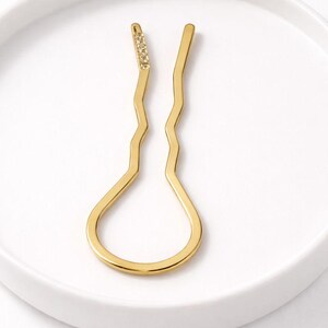 May include: A gold-coloured hair pin with a distinctive, wavy design. The top of the pin features a row of small, sparkling embellishments. The pin is set against a white background, highlighting its elegant form.