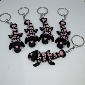May include: Keychains featuring a black and pink alligator design. The alligator figures are decorated with pink hearts and attached to silver key rings. The keychains are displayed on a white surface.