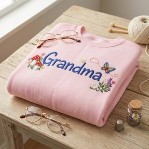 May include: A light pink sweatshirt embroidered with the word "Grandma" in blue script. The sweatshirt features floral and butterfly embroidery. Two pairs of gold-rimmed glasses and a small jar of flowers are also present.
