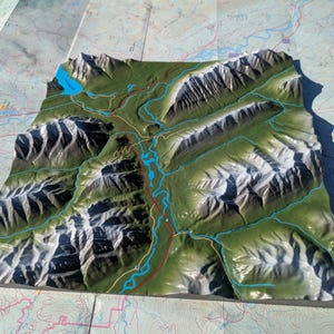 Peut inclure: Une maquette de carte topographique 3D représentant des chaînes de montagnes détaillées avec des sommets enneigés, des vallées vertes et des lacs et rivières bleus. Le modèle est posé sur une carte papier détaillée.